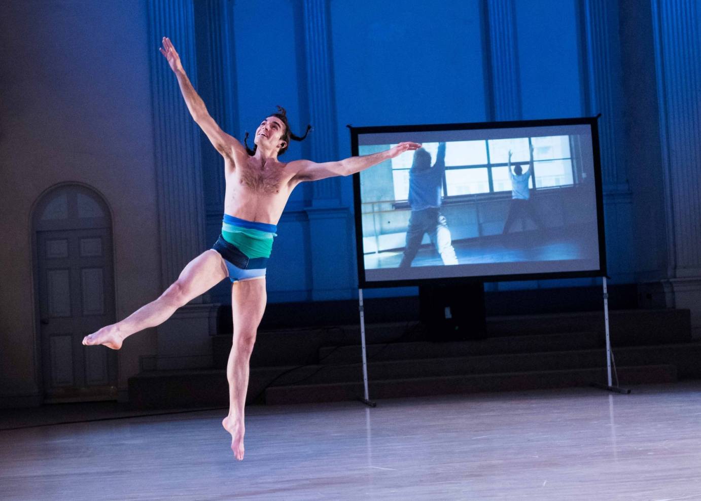 Silas Riener smiling in a leotard while rehearsal footage of him and Adrian Danchig-Waring is projected in the background